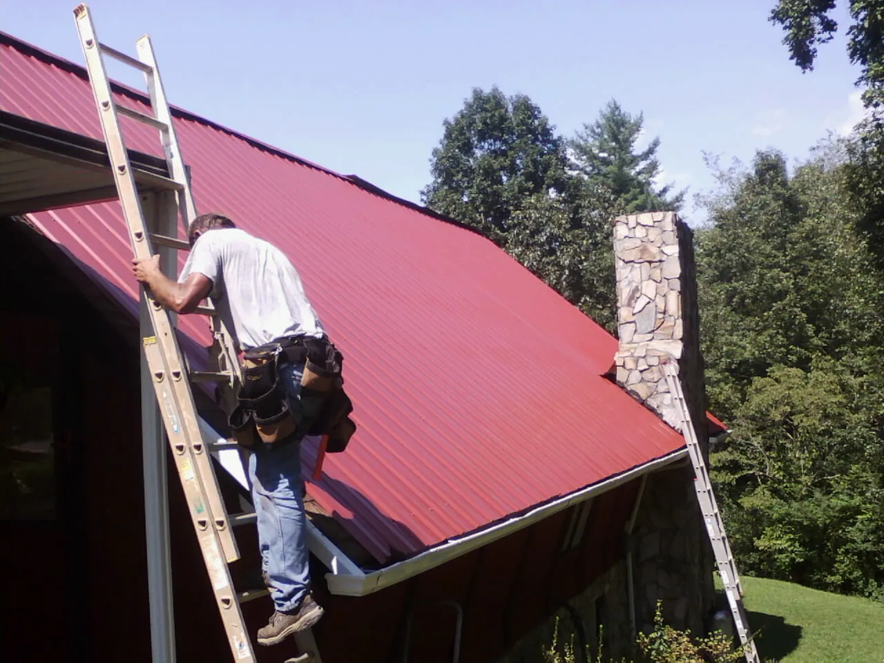 Roofer on a ladder beside a red metal roof and stone chimney — Soffit & Fascia Repair by Roofing Brothers in Crawfordsville